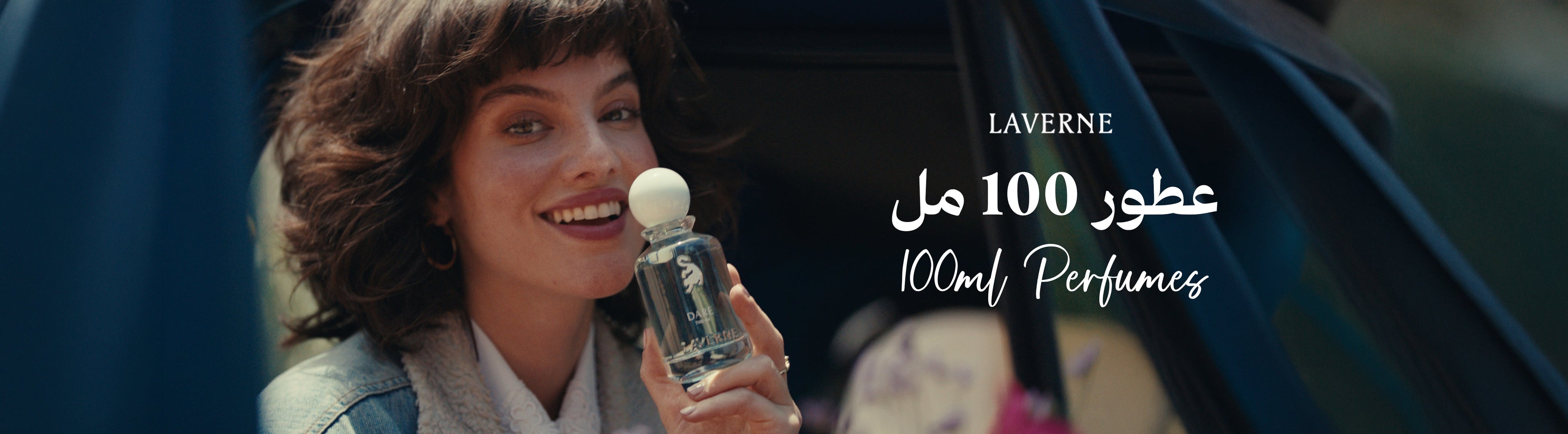 Close-up of a smiling woman with short brown hair inside a car, holding a transparent 100ml Laverne perfume bottle. The background includes text in both Arabic and English: "عطور 100 مل / 100ml Perfumes".